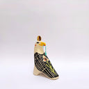 A stylized ceramic bird figurine in cream, black, and yellow, with a small red blush dot on its cheek and a green hanging tassel in its beak, photographed against a plain light gray background.