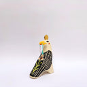 A stylized ceramic bird figurine in cream, black, and yellow, with a small red blush dot on its cheek and a green hanging tassel in its beak, photographed against a plain light gray background.