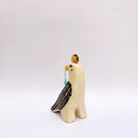 A stylized ceramic bird figurine in cream, black, and yellow, with a small red blush dot on its cheek and a green hanging tassel in its beak, photographed against a plain light gray background.