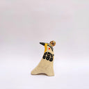 Cream colored ceramic sculpture of a bird, sitting in profile, with black and yellow feather detailings. It has a red cheek and a gold coin atop its head.
