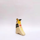 Cream colored ceramic sculpture of a bird, sitting in profile, with black and yellow feather detailings. It has a red cheek and a gold coin atop its head.