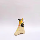 Cream colored ceramic sculpture of a bird, sitting in profile, with black and yellow feather detailings. It has a red cheek and a gold coin atop its head.