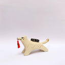 A ceramic sculpture shaped like a wolf, cream colored with gold color detailings. It carries a red tassel in its mouth and has a small wrapped doll-like object on its back.