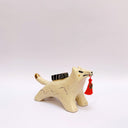 A ceramic sculpture shaped like a wolf, cream colored with gold color detailings. It carries a red tassel in its mouth and has a small wrapped doll-like object on its back.