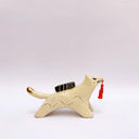 A ceramic sculpture shaped like a wolf, cream colored with gold color detailings. It carries a red tassel in its mouth and has a small wrapped doll-like object on its back.