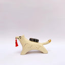 A ceramic sculpture shaped like a wolf, cream colored with gold color detailings. It carries a red tassel in its mouth and has a small wrapped doll-like object on its back.
