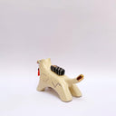 A ceramic sculpture shaped like a wolf, cream colored with gold color detailings. It carries a red tassel in its mouth and has a small wrapped doll-like object on its back.