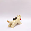 A ceramic sculpture shaped like a wolf, cream colored with gold color detailings. It carries a red tassel in its mouth and has a small wrapped doll-like object on its back.
