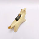 A ceramic sculpture shaped like a wolf, cream colored with gold color detailings. It carries a red tassel in its mouth and has a small wrapped doll-like object on its back.