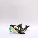 A ceramic orca sculpture with a black and cream body, decorated with cream sgraffito line art and flowers, and a turquoise tassel ornament on its side, set against a plain white background.