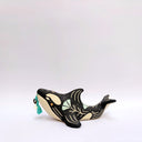 A ceramic orca sculpture with a black and cream body, decorated with cream sgraffito line art and flowers, and a turquoise tassel ornament on its side, set against a plain white background.