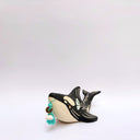 A ceramic orca sculpture with a black and cream body, decorated with cream sgraffito line art and flowers, and a turquoise tassel ornament on its side, set against a plain white background.