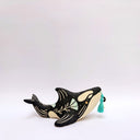 A ceramic orca sculpture with a black and cream body, decorated with cream sgraffito line art and flowers, and a turquoise tassel ornament on its side, set against a plain white background.