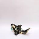 A ceramic orca sculpture with a black and cream body, decorated with cream sgraffito line art and flowers, and a turquoise tassel ornament on its side, set against a plain white background.