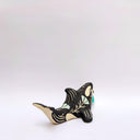 A ceramic orca sculpture with a black and cream body, decorated with cream sgraffito line art and flowers, and a turquoise tassel ornament on its side, set against a plain white background.