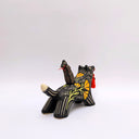 Ceramic sculpture of a wolf, black with cream colored sgraffito fur textures. It has a poppy flower design on its side and on its back is a doll charm of a woman, with a tiny wolf's mask atop her head. The wolf has a red tassel in its mouth.