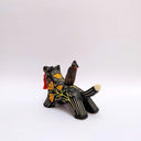 Ceramic sculpture of a wolf, black with cream colored sgraffito fur textures. It has a poppy flower design on its side and on its back is a doll charm of a woman, with a tiny wolf's mask atop her head. The wolf has a red tassel in its mouth.