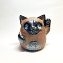 Ceramic sculpture of a cute, brown cat with one paw on its cheek and the other raised up. It has a rounded bottom without legs and black spots on its muzzle, paws, stomach and ears.