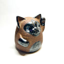 Ceramic sculpture of a cute, brown cat with one paw on its cheek and the other raised up. It has a rounded bottom without legs and black spots on its muzzle, paws, stomach and ears.