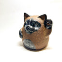 Ceramic sculpture of a cute, brown cat with one paw on its cheek and the other raised up. It has a rounded bottom without legs and black spots on its muzzle, paws, stomach and ears.