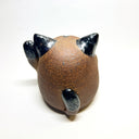Ceramic sculpture of a cute, brown cat with one paw on its cheek and the other raised up. It has a rounded bottom without legs and black spots on its muzzle, paws, stomach and ears.