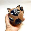 Ceramic sculpture of a cute, brown cat with one paw on its cheek and the other raised up. It has a rounded bottom without legs and black spots on its muzzle, paws, stomach and ears.