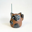 Ceramic sculpture of a cute, brown cat with one paw on its cheek and the other raised up. It has a rounded bottom without legs and black spots on its muzzle, paws, stomach and ears.