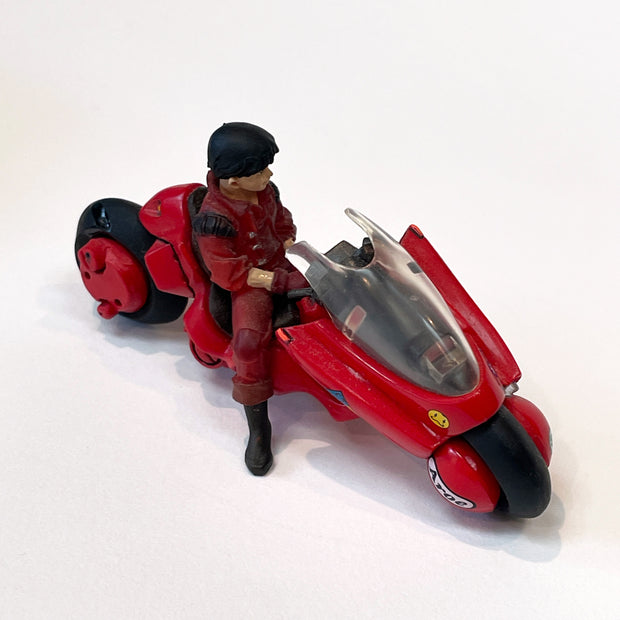 Vinyl figure of Kaneda from Akira, a young boy, wearing an all red leather outfit and sitting on the famous bike from Akira. He leans back in the seat with his hands in his lap.