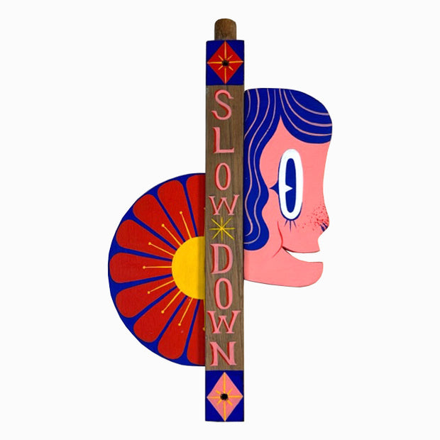 Die cut painted wooden sculpture of a vertical post that reads "Slow Down" in stylized letters. On the right side of the post is a cartoon woman's face popping out and on the left is half a red flower.
