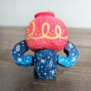 Whittled sculpture of a character with a  red vessel pouring out patterned water as its head and its body made out of the same water patterning. It's head has a sad expression and gold colored accents and it sits and holds its head in its hands. 