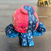 Whittled sculpture of a character with a  red vessel pouring out patterned water as its head and its body made out of the same water patterning. It's head has a sad expression and gold colored accents and it sits and holds its head in its hands. 