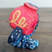 Whittled sculpture of a character with a  red vessel pouring out patterned water as its head and its body made out of the same water patterning. It's head has a sad expression and gold colored accents and it sits and holds its head in its hands. 