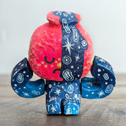 Whittled sculpture of a character with a  red vessel pouring out patterned water as its head and its body made out of the same water patterning. It's head has a sad expression and gold colored accents and it sits and holds its head in its hands. 