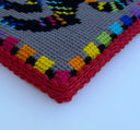 A close-up of a crocheted or knitted textile corner showing a gray base with a multicolored geometric pattern and a red textured border on a white background.