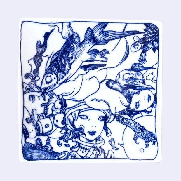 White porcelain plate with stark blue line illustration of 3 girls with robotic helmets, seen only from the neck up. A fish is positioned across the plate with a bird below it.