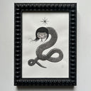 Framed artwork of a stylized illustration with a snake with 2 pairs of eyes and long black hair.