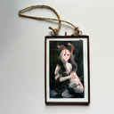 A framed artwork featuring a stylized painting of a woman crouched down, blanketed with her own hair that looks like smoke. Her eyes are pulled out of her head, attached by a red tendon.