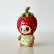 Small ceramic sculpture of a character wearing a giant strawberry around their head. They sit with a tiny body.