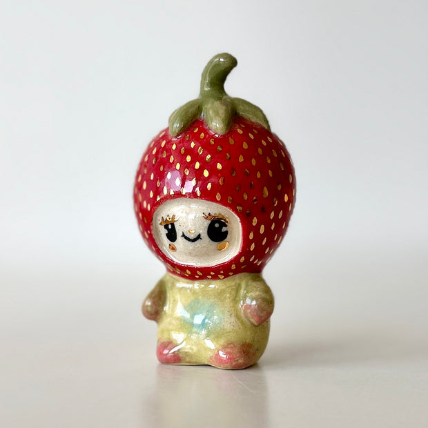 Small ceramic sculpture of a character wearing a giant strawberry around their head. They sit with a tiny body.