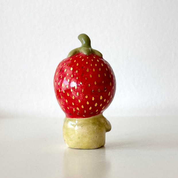 Small ceramic sculpture of a character wearing a giant strawberry around their head. They sit with a tiny body.