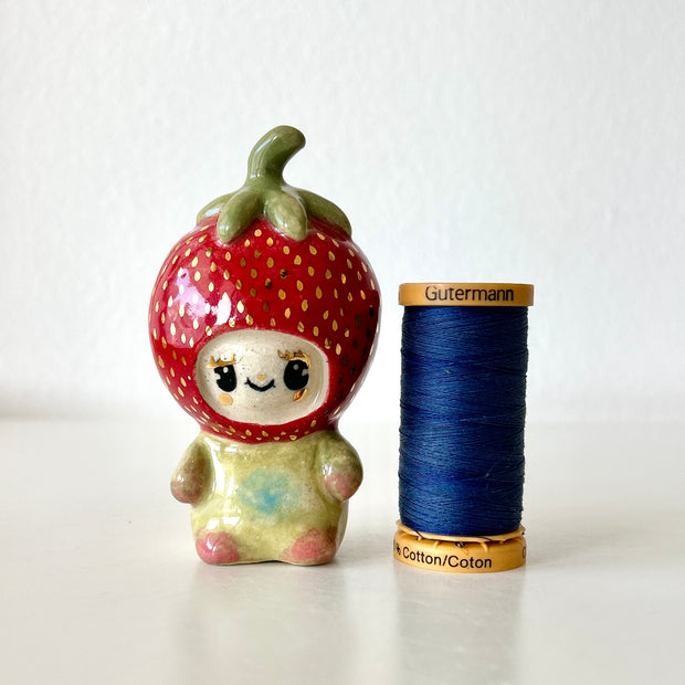Small ceramic sculpture of a character wearing a giant strawberry around their head. They sit with a tiny body next to a spool of thread for size reference.