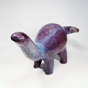 Purple and blue glossy ceramic sculpture of a long neck dinosaur with a simplistic, cute face.