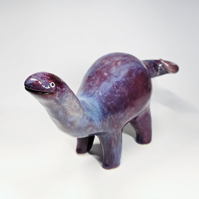Purple and blue glossy ceramic sculpture of a long neck dinosaur with a simplistic, cute face.