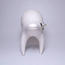 White ceramic sculpture of a quadruped creature with a large humped body, curved tall legs and a long neck. It has cracks running through its body and inside of it grows flowers. 