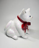 White cat-shaped ceramic sculpture with pink glitter specks, wearing a sparkly red bow.