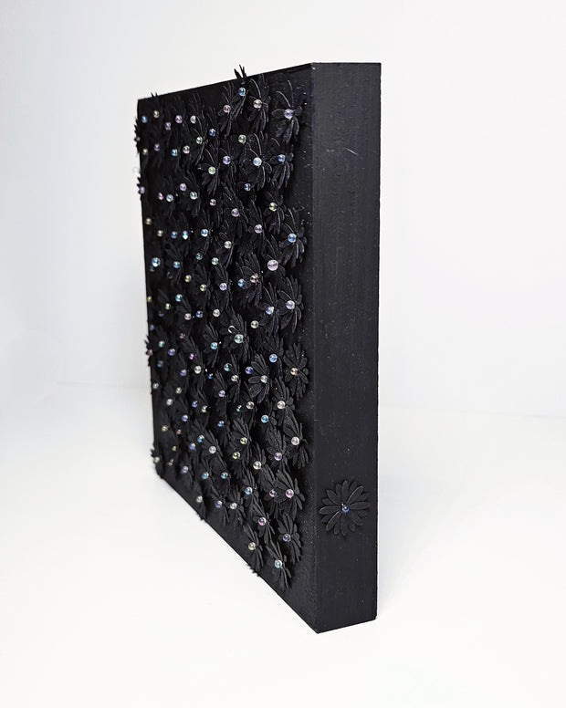 Black wooden panel covered in small glittery black flowers cut out of paper and adhered, coating most of the panel's surface. Each flower has a small gem as the center.