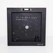 Back of a black wooden panel with small black glittery flowers cut out of paper and adhered. A small black character with a rounded body and small wings sits on the inner ledge of the pane.