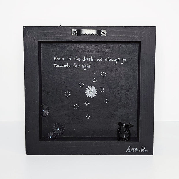 Back of a black wooden panel with small black glittery flowers cut out of paper and adhered. A small black character with a rounded body and small wings sits on the inner ledge of the pane.