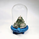 Sculpture encased in a glass tall dome with a blue clay base shaped like a turtle. Within the dome is a rocky hill landscape with moss and tiny houses.