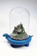 Sculpture encased in a glass tall dome with a blue clay base shaped like a turtle. Within the dome is a rocky hill landscape with moss and tiny houses.